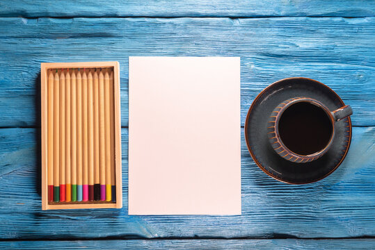 Blank paper page with copy space, colorful pencils and black coffee cup on the blue desk background.