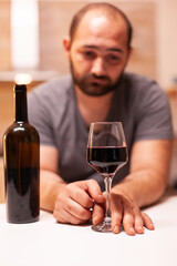 Glass of wine in focus with depressed young man in the background. Unhappy person disease and anxiety feeling exhausted with having alcoholism problems.