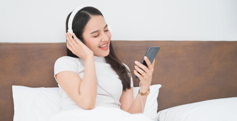 Beautiful young asian woman with headphones relaxing on the bed. She is listening to music using smartphone or tablet. Chill out and leisure concept
