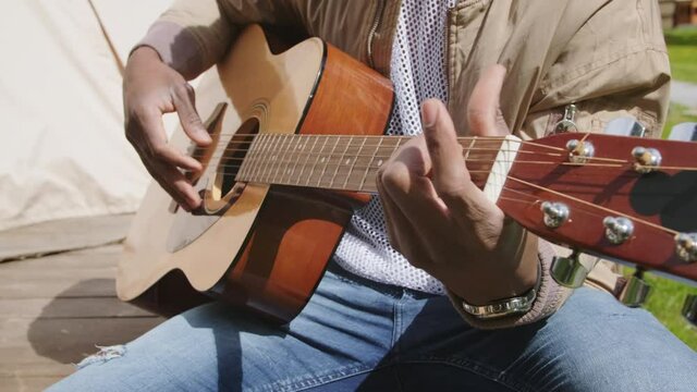 Closeup of unrecognizable African American man fingers touching strings of wooden guitar playing melody sitting outside enjoying summertime