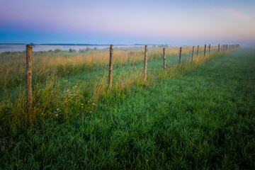 Obraz premium Morning fog in the meadows during sunrise in the countryside. Rural landscape with a fog on the geen field. the fence stretching into the distance.