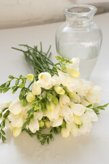 Obraz na płótnie Canvas Vertical close up of white freesia flowers next to glass vase (selective focus)