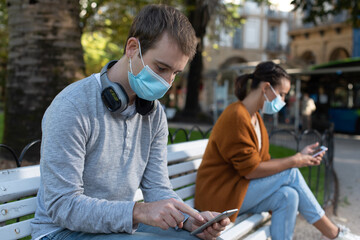 Young man and woman sitting on a bench checking their phones while keeping social distance during Covid-19 pandemic. Couple wearing a face mask.
