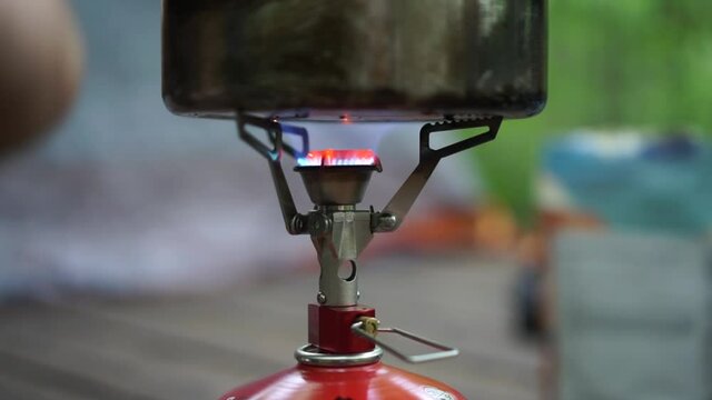 Burning Camping Stove Boiling Water with Man Placing Food Packet Down In Background.