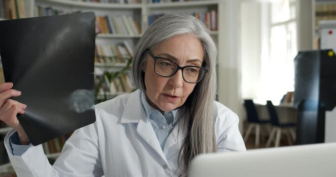 Close up of female doctor showing x ray photo while having online medical consultation in office. Mature woman in glasses and white gown using laptop while communicating with patient