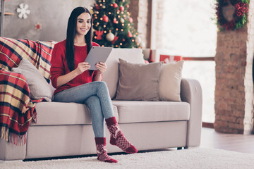 Photo of cute young lady sit couch hold tablet toothy smiling wear red sweater denim jeans socks in decorated living room indoors