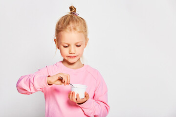 Cute little girl eating tasty yogurt on gray background. 4-5 year old kid posing in studio in a pink sweatshirt. Space for text