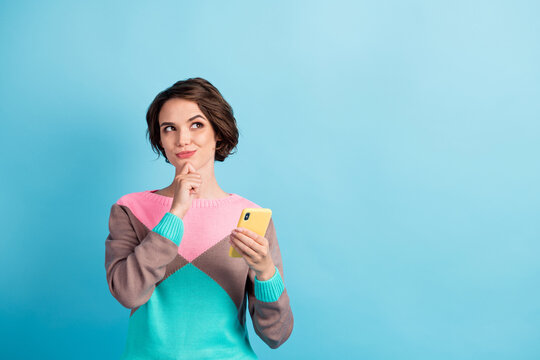 Photo portrait of curious woman holding phone in one hand touching chin with finger looking at blank space isolated on pastel light blue colored background