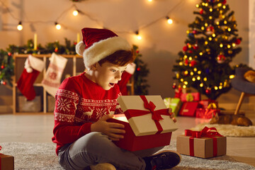 Obraz na płótnie Canvas Boy in Santa holiday hat and red festive sweater feeling surprised opening holiday Christmas gift box