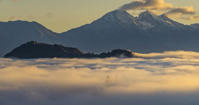 Time lapse low altitude inversion clouds rolling in Alpine valley, Slovenia. Saint Thomas church tower visible above the fog. Alpine mountains in background. Right truck parallel moving, wide angle