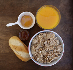 Breakfast table scene with a bowl of corn flakes, apricot jam, brioche, honey and orange juice on a wooden table