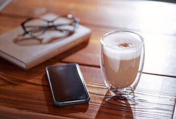 Coffee break. Glass of cappuccino, smartphone and book on wooden table.