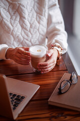 Working and relaxing at cafe. Close up of female hands holding cup of cappuccino coffee on wooden tavble with laptop computer.