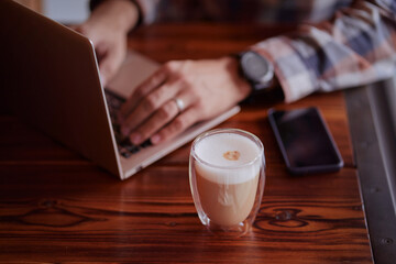 Coffee break. Cup of cappuccino on working table with a man typing on a laptop.