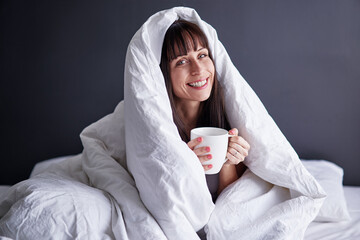 Time for myself. Comfort and relaxation. Pretty young woman drinking tea while sitting on her bed at home covering by blanket.