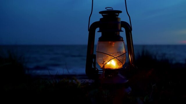 Old classic oil lantern burning with an orange flame by the ocean at dusk. Blurred background.