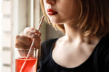 A young girl with bright red lipstick drinks a cocktail from a glass from a paper tube. The concept of ecology and environmental protection.
