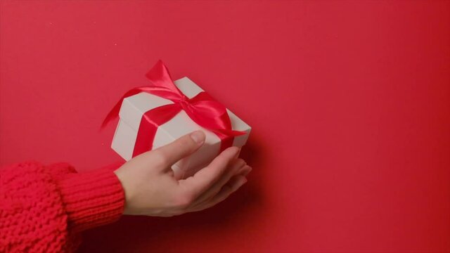 A gift in a box with a red ribbon falls from above into the girl's hands. Slow motion. Women's hands in a red knitted sweater receive a gift for the holiday. Red background.