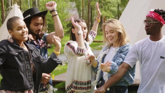 Medium footage of cheerful multi-ethnic group of people dancing and chilling at campsite with tents and forest in background