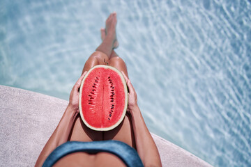 Concept of diet, spa and cosmetics. Enjoying tropical fruits, suntan and vacation. Young slim woman in green swimsuit sitting near swimming pool holding watermelon.