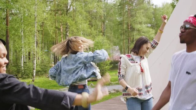 Joyful multi-ethnic male and female friends putting hands in air simultaneously and dancing outdoors spending day in campsite among trees and tents