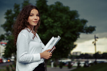 Vertical portrait of young lovely curly woman standing outside with documents and looking at the camera, smiling. Lady dressed in white shirt and black skirt going to the job in early morning.