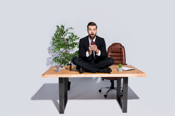 Full body photo of handsome business guy hold telephone hands sitting table legs crossed read bad fake shock news wear blazer shirt pants shoes suit isolated grey background