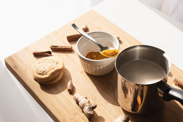 Cutting board with curcuma powder in white bowl,  pot with almond milk, cinnamon sticks and row turmeric roots. Preparation steps of homemade curcuma latte.