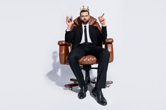 Full body low below angle view photo of handsome business guy sit chair hold rum glass cuban cigar chief wear crown black blazer pants tie shirt shoes suit isolated grey background