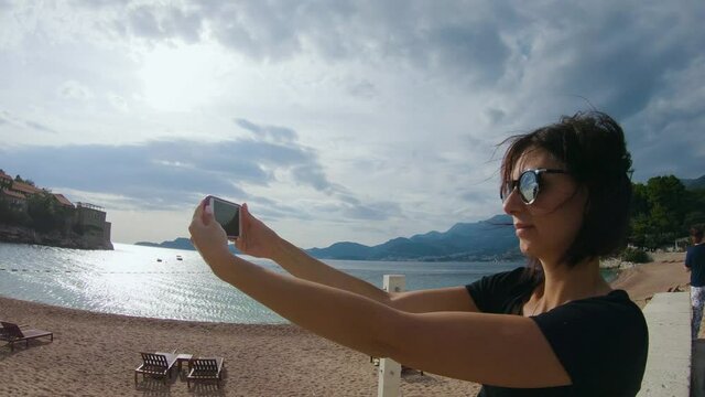 Side view. Pretty young woman in sunglasses takes selfie on her smartphone on the island Sveti Stefan in Montenegro