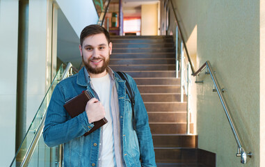 Student on stairs. Portrait caucasian smiling handsome bearded man with backpack notebooks in reopen university campus hallway. Freelancer in modern coworking office