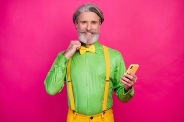 Photo of funny white haired grandpa hold telephone look mirror fix elegant bow tie metrosexual wear green shirt yellow suspenders pants isolated shine bright pink color background