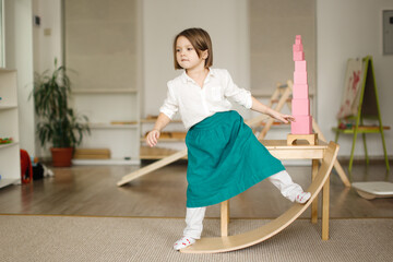 child girl in a linen apron sways on balancing board, developing sensory and sports activities in montessori and earlier child development, children's independence - obrazy, fototapety, plakaty