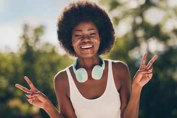 Portrait of positive dark skin girl enjoy summer forest park walk make v-sign wear white singlet