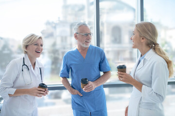 Group of smiling doctors drinking coffee next to the window, having friendly conversation