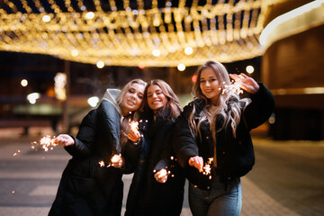Fototapeta na wymiar Caucasian young girls in jackets with sparklers on the street, a cheerful company of girls and celebration, new year and christmas