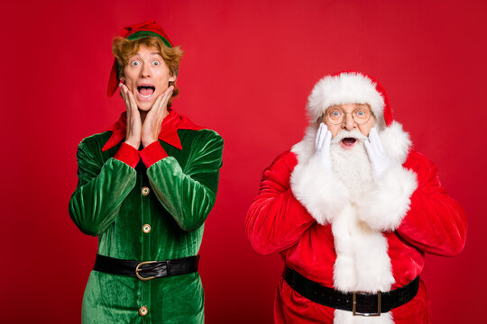 Photo of two men santa claus elf open mouth arms cheeks wear x-mas costume coat headwear specs isolated red color background