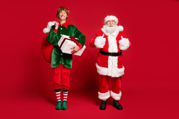 Full length photo of two men santa elf hold bag box raise thumb up wear x-mas costume coat cap...