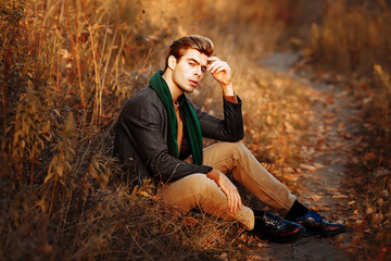 a man, a model in a gray jacket, a green scarf and beige pants and a sweater sits on the grass in...