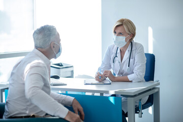 Blonde female doctor in mask taking notes, consulting gray-haired male patient