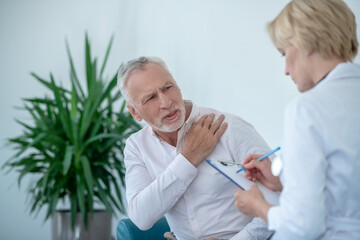 Fototapeta premium Gray-haired male patient suffering from shoulder pain, female doctor taking notes
