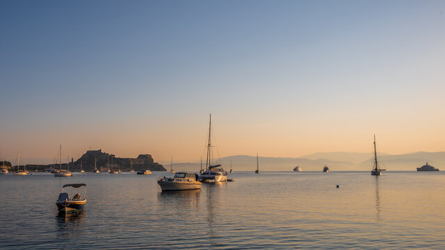 Corfu Town harbor front, at sunrise. Multiple small boats can be seen in the sea.