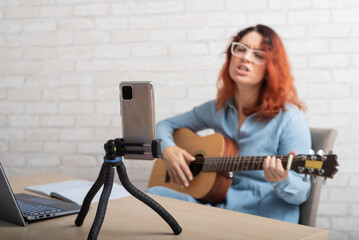 Fototapeta na wymiar A woman is broadcasting online on her phone at home. The girl sings and plays the guitar live