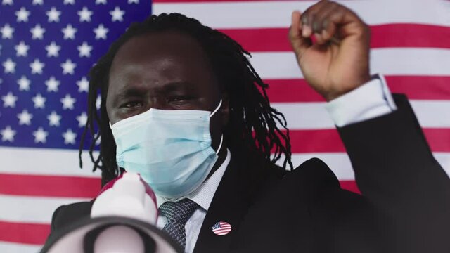 Black Man Protester Wearing Face Mask Holding A Megaphone Yelling With Clenched Fist Standing Against United States Flag Background - Protest Amid Covid-19 Pandemic - Closeup Shot