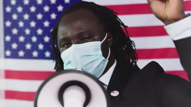 African-American Man Wearing Face Mask Speaking Loud Into Megaphone Protesting While Raising Arm Clenching Fist In The Air With USA Flag Background - Closeup Shot