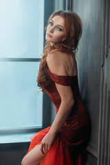 Portrait Young beautiful woman in an elegant luxury red dress stands near window in vintage dark...