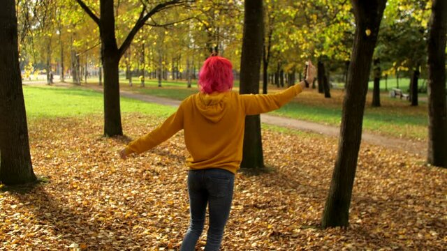 Young smiling hipster woman with bright pink hair is having fun, spinning, posing and taking selfie self portrait photos on smartphone. Walk in the autumn park. Fallen yellow leaves lie on the ground.