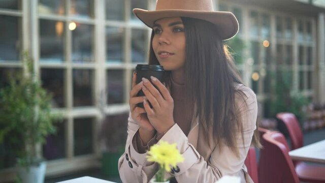 portrait of a young beautiful woman sitting in a cafe holding a disposable glass with hot coffee or tea. High quality 4k footage