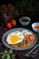 Mie Goreng or fried noodles, typically Indonesian fried noodles topping with sunny side up egg, cucumber sliced, and tomato. Served with sambal as condiment. Selective focus on dark wooden background.