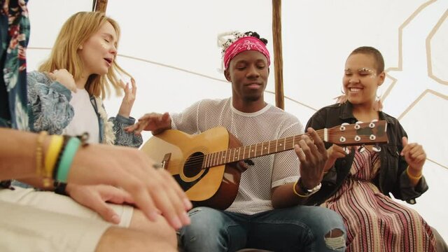 Multi-ethnic friends having fun together sitting in tent and singing and listening to music played by talented African American musician with dreadlocks